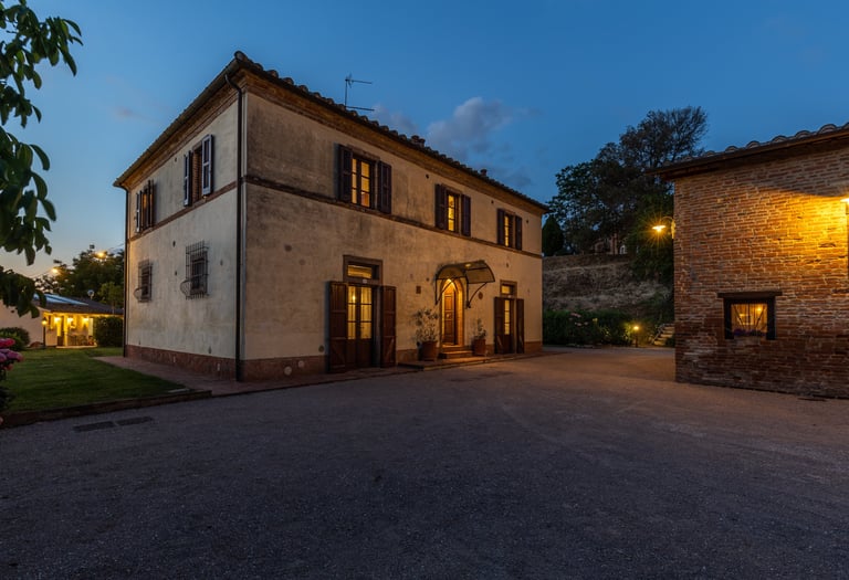 A historic Tuscan villa exterior at dusk with warm glowing lights and a gravel courtyard.