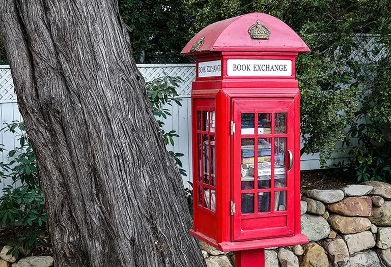 Red British phone-booth style Book Exchange box filled with books next to a large tree trunk.
