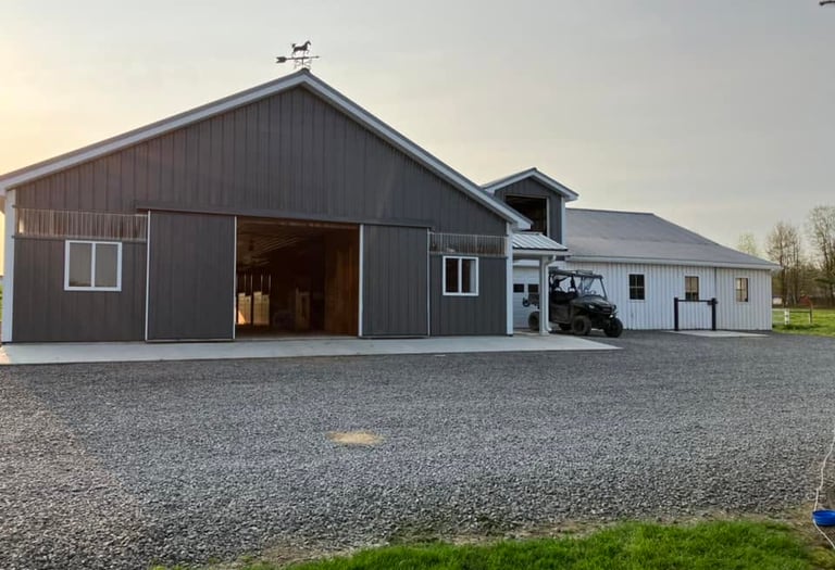 A modern charcoal grey metal horse barn with sliding doors and a white farm building on a gravel driveway.
