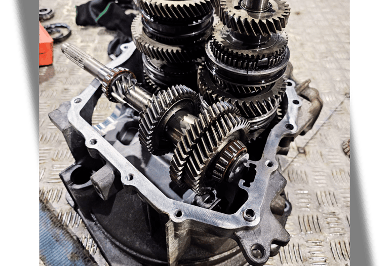 Close-up of internal metal gears and shafts inside a disassembled car transmission gearbox on a workshop floor.