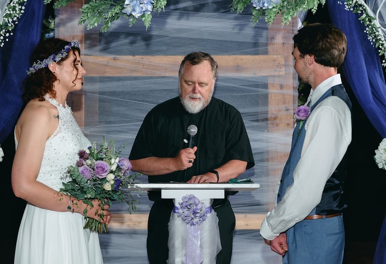 a groom and a bride standing across each other and a priest standing in between them giving a speech