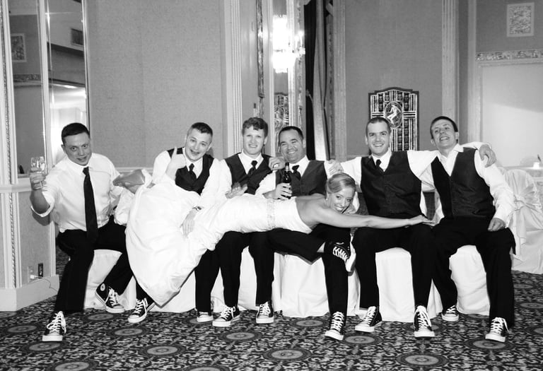 6 men in suits sitting on white chairs with a bride lying on them
