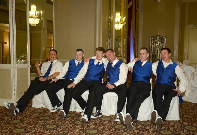a group of men in suits and blue ties and blue vests sitting on white chairs at a hotel ball room