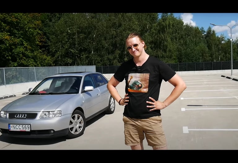 Youtuberis su juodais marškinėliais šalia sidabrinio Audi A3 saulėtoje aikštelėje.