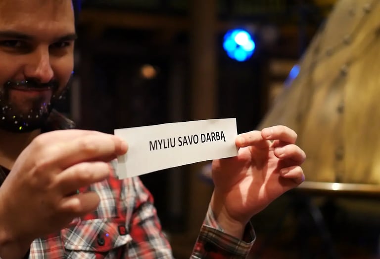 Vyras su blizgančia barzda, įmonės vakarėlyje, laiko lapą su užrašu „MYLIU SAVO DARBĄ"