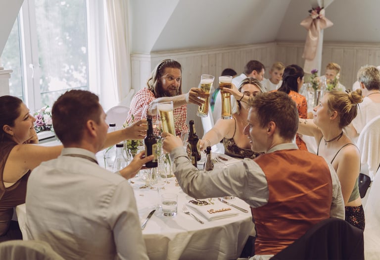 Hochzeitsgäste stoßen fröhlich mit Biergläsern am Tisch an. Authentische Eventfotografie von Just Perspectives.