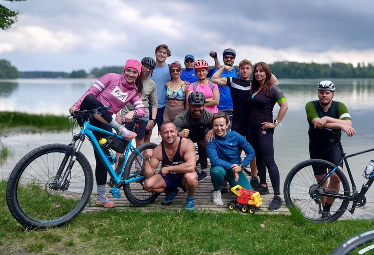 Uśmiechnięta grupa osób pozuje do zdjęcia z rowerami na tle jeziora.
