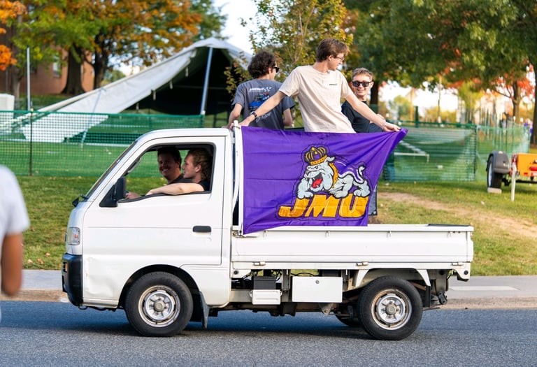 Luke Gilmore riding on the back of a Kei truck with a JMU banner on it.