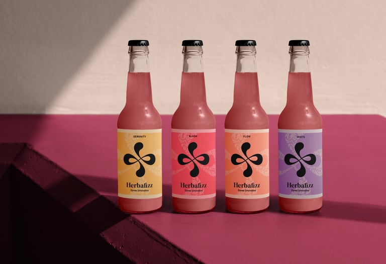 Four glass bottles of Herbafizz sparkling botanical lemonade with colorful labels in natural sunlight.