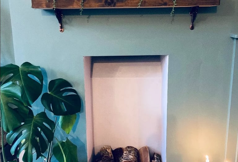 Rustic indoor fireplace featuring a succulent wooden mantle shelf, monstera plant, and lit candles.