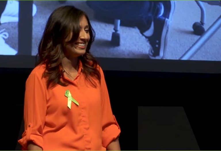A woman in an orange shirt smiles on stage, wearing a green awareness ribbon, conveying positivity.