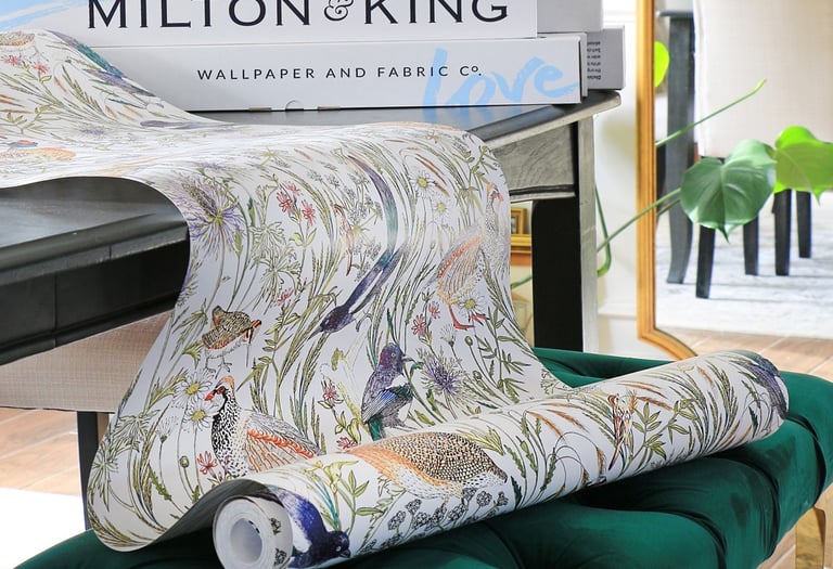 Milton and King floral bird wallpaper roll draped over a green velvet ottoman in a bright room.