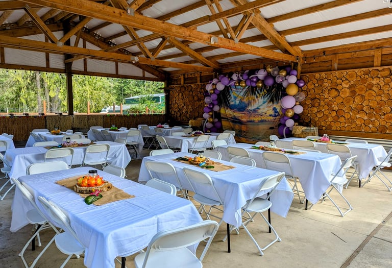 Rustic outdoor event venue featuring decorated banquet tables under a wooden pavilion with a tropical balloon arch.
