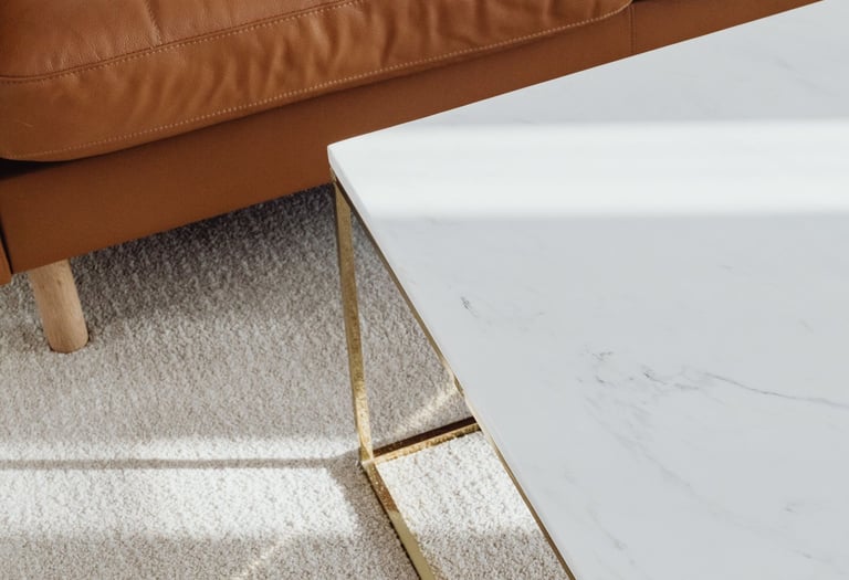 Modern white marble coffee table with gold legs sitting on a plush rug next to a tan leather sofa.