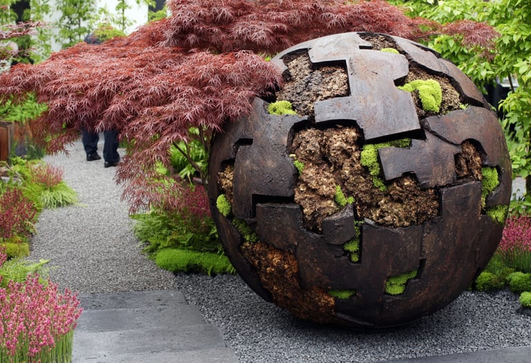 Rustic metal sphere garden sculpture nestled among red Japanese maple trees and lush green moss.