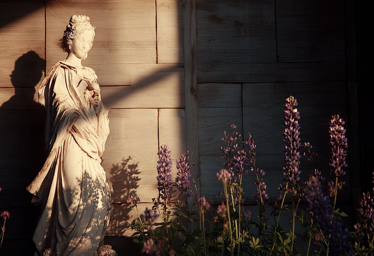 Elegant female garden statue standing near purple lupine flowers at sunset with warm shadows.