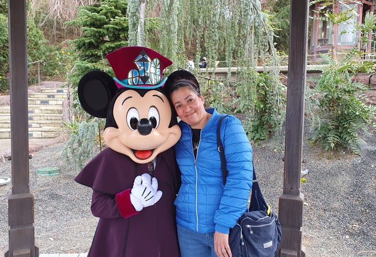 A smiling woman in a blue jacket poses with Mickey Mouse in a purple costume at a Disney park.