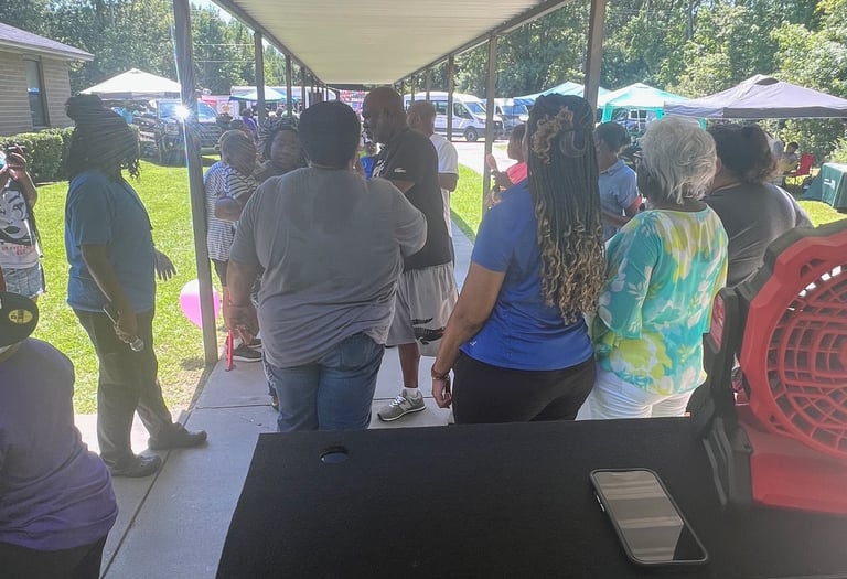 A DJ booth setup at an outdoor community event with people gathered under a long canopy.