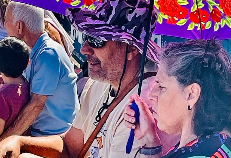 A man in a purple hat and a woman sit under a floral purple sun umbrella at an outdoor event.