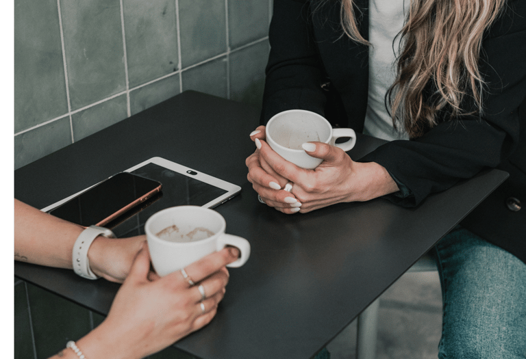 Two women holding coffee mugs while sitting at a cafe table with a smartphone and tablet.