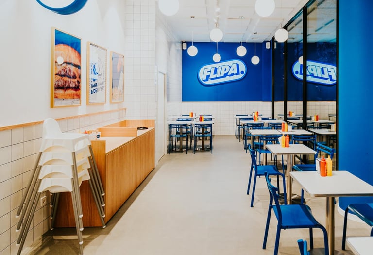 Vista de l’interior de Flipa Burger amb logotip a la paret blava, decoració retro i ambient lluminós