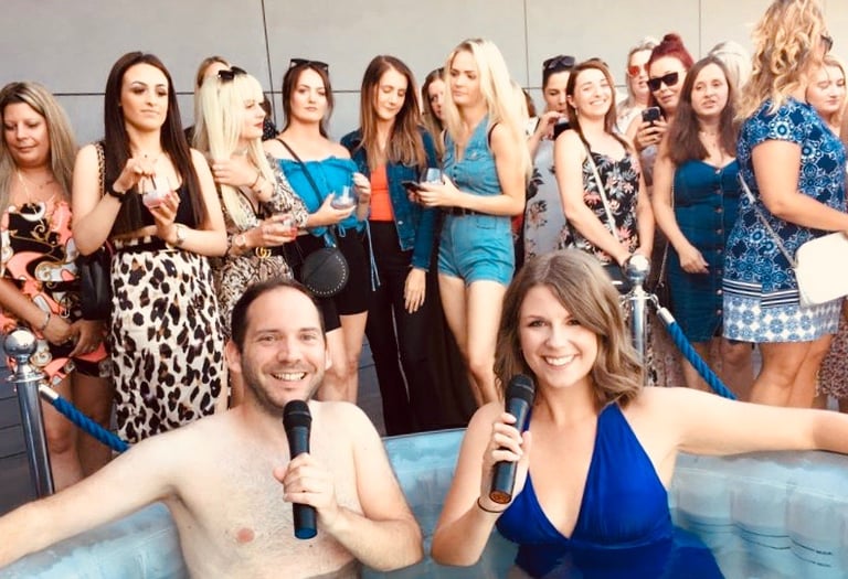 Alex Duffy and Ellie Brennan are in a hot tub with microphones, with audience behind.