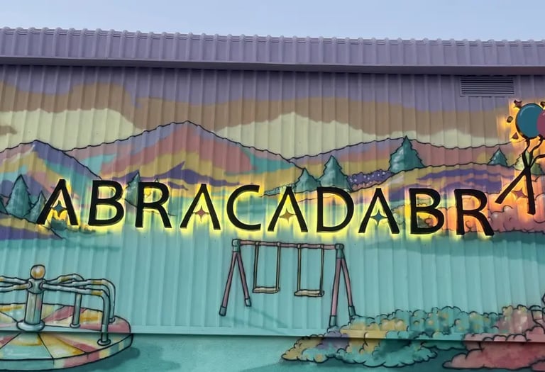 Backlit Abracadabra sign on a colorful playground mural with painted mountains and balloons.