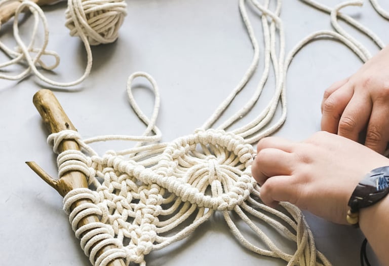 Manos elaborando un colgante de pared de macramé DIY usando cordón de algodón blanco con una rama de madera.