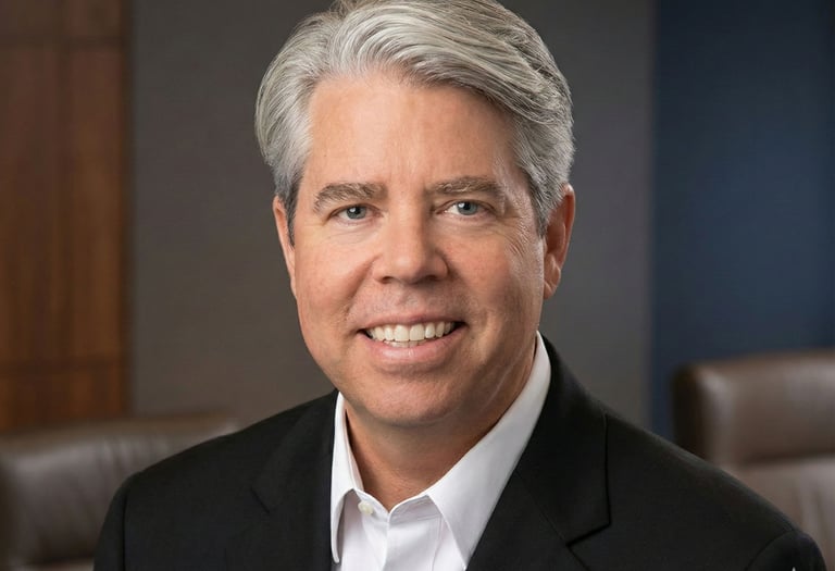 Professional headshot of a smiling male executive with grey hair in a black suit jacket and white shirt.