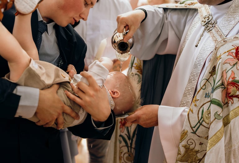 Krikštynų fotografija. Vyksta berniuko krikštas. Kunigas lieja vandenį ant vaiko kaktos.