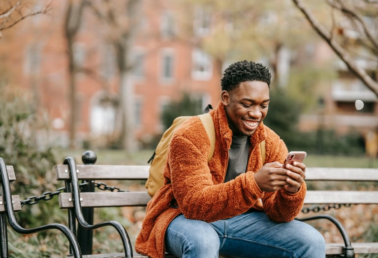 A smiling young man in a fleece jacket using a smartphone while sitting on a park bench.