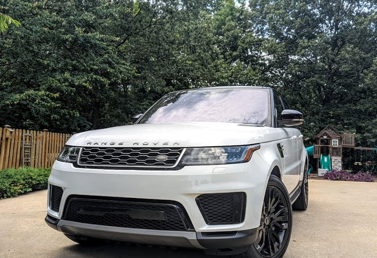 White Land Rover Range Rover Sport with black rims parked on a driveway near green trees.