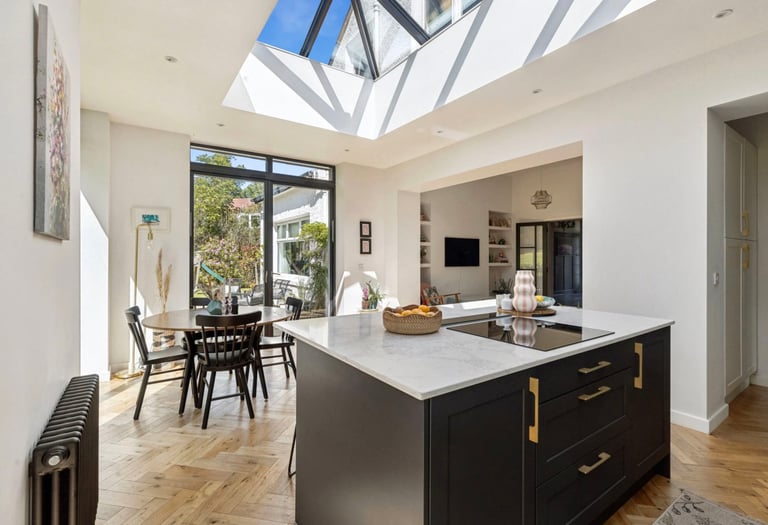 Modern open plan kitchen featuring a dark island with marble countertop and a large skylight roof lantern.