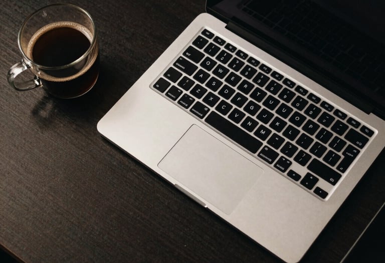 Top-down view of a designer's desk with a high-end laptop, glass coffee mug, and neon-lit keyboard in a dark North American creative studio.