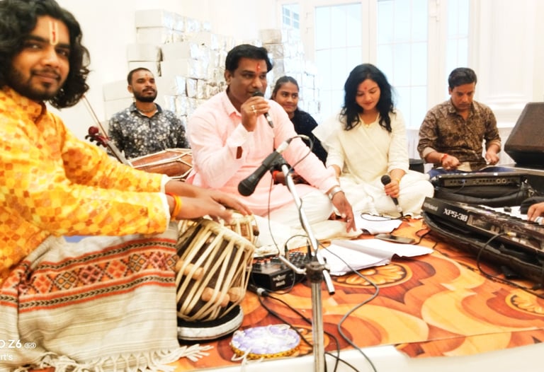 Indian musicians performing a devotional bhajan with tabla drums, harmonium, and vocal microphones.