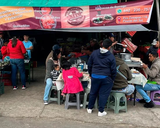 Puesto de Carnitas y Barbacoa NITO con clientes comiendo en mesas bajo una lona en un mercado