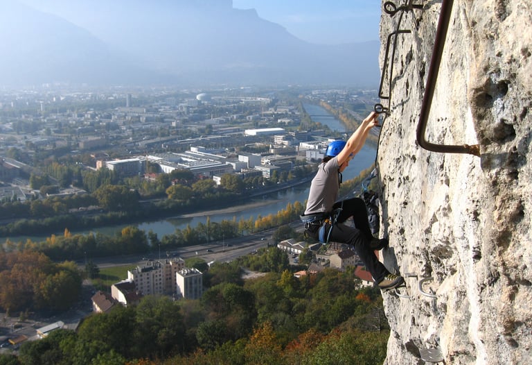 A mountain climber climbs a steep via ferrata route overlooking a scenic city valley and river.