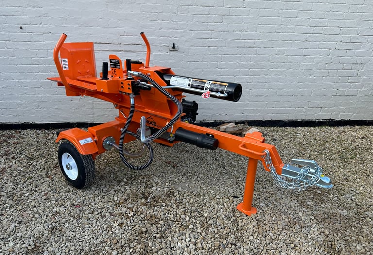 Heavy duty orange hydraulic log splitter machine parked on gravel against a white brick wall.