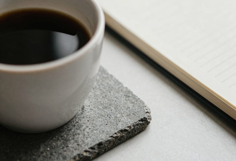 Close-up of a cup of coffee on a grey stone coaster next to a closed notebook, neutral colors, representing a quiet moment of introspection.