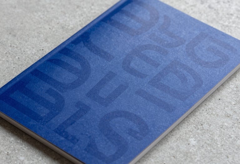 Close-up photography of a high-quality printed marketing brochure featuring bold typography and deep dark blue colors, lying on a light grey concrete surface.