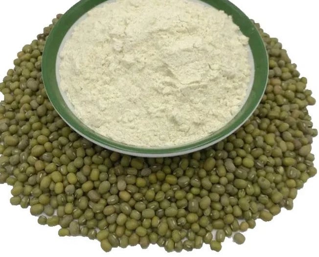 A green bowl of fine white mung bean flour sitting on a pile of raw green mung beans.