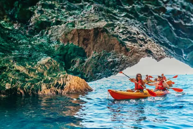 Kayakers in red life jackets paddling through a sea cave into turquoise Costa Brava water