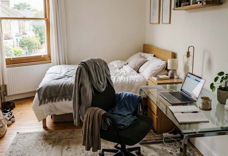 European bedroom with a chair covered in clothes, a desk with a laptop and mug, and a slightly unmade bed