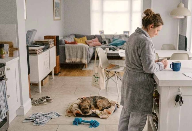 Woman in a dressing gown standing in a softly lit European home with a small dog resting nearby and everyday clutter around