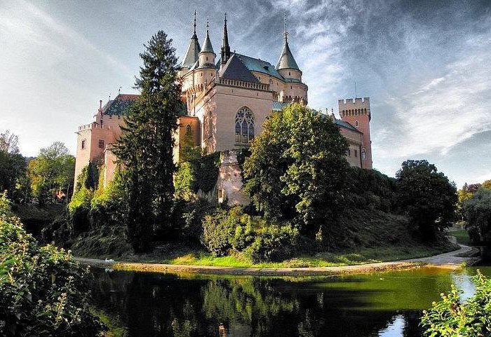 Bojnice Castle in Slovakia reflecting in a lake surrounded by lush green trees under a cloudy sky.