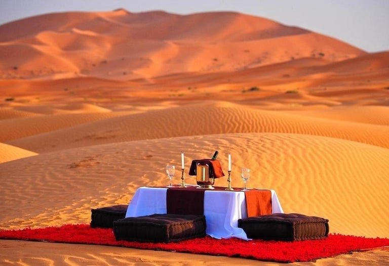 A romantic luxury dinner setup for two on a red rug among the Sahara Desert sand dunes at sunset.
