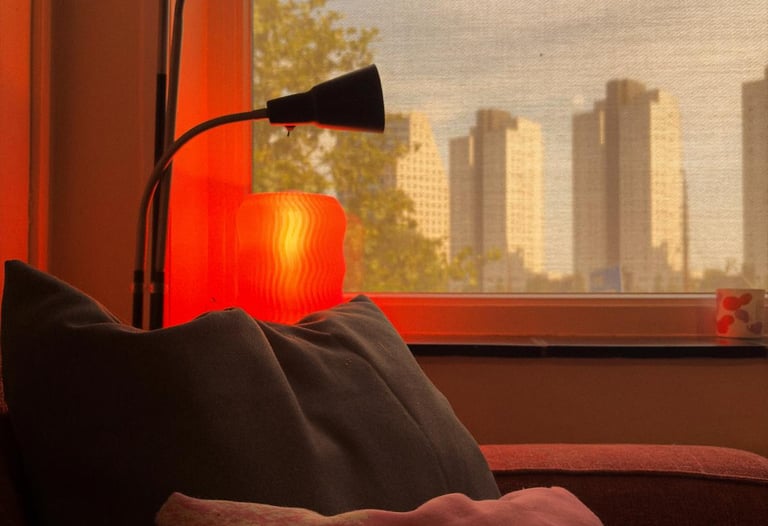 Cozy living room corner with a red glowing lamp, plush pillows, and a window view of city skyscrapers at sunset.