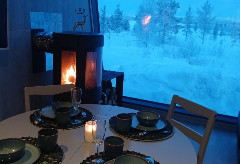 Cozy dinner table set for four by a wood-burning fireplace with a snowy winter landscape view.