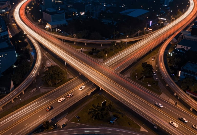 Drone photography of a complex highway interchange at night, with light trails from cars creating vibrant orange and white lines against a dark blue background.
