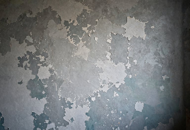 A detailed close-up of a weathered North American interior wall on a film set. The surface shows layers of peeling silver-grey paint and dark teal undertones, masterfully aged by a scenic artist. Soft, moody side-lighting highlights the texture and depth of the plaster work.
